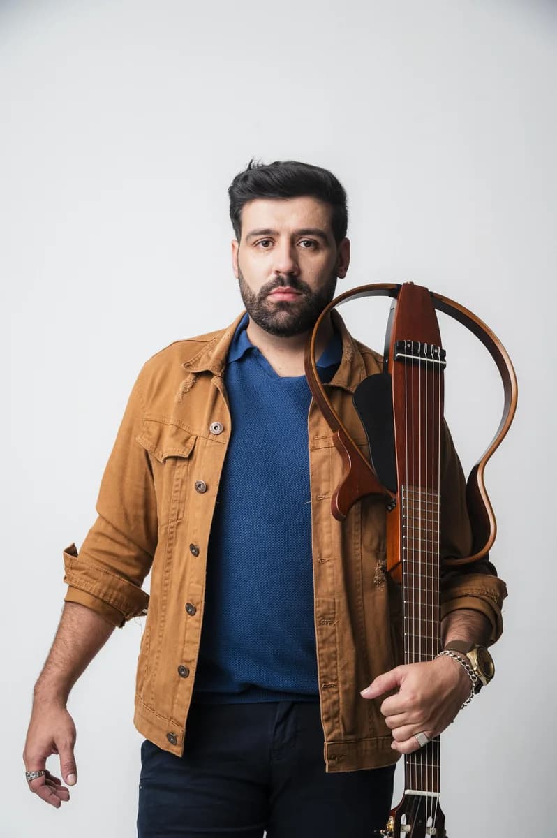 Geovani Medeiros em retrato de estúdio, de jaqueta caramelo, segurando o violão ao lado do corpo