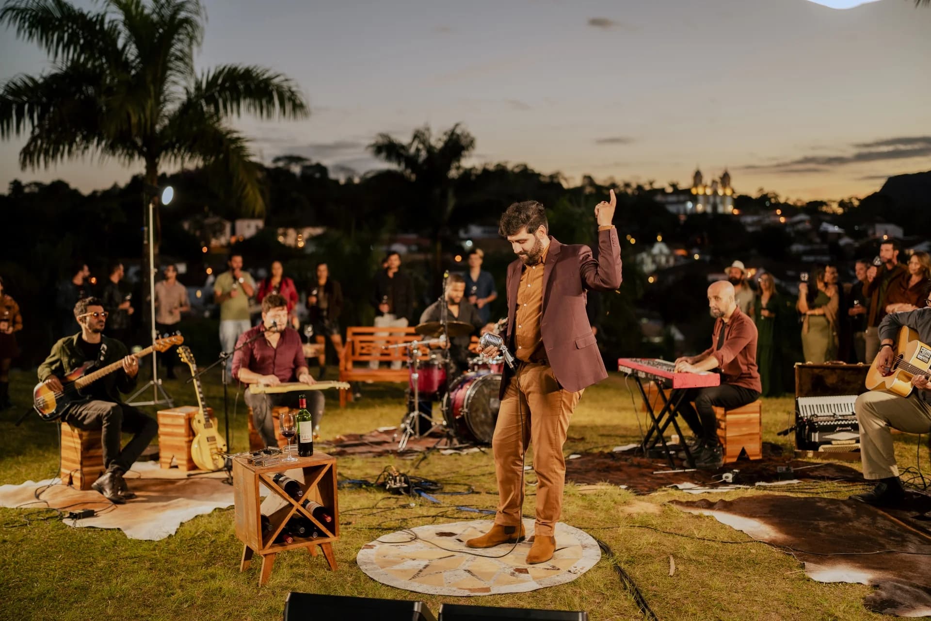 Geovani Medeiros de braço erguido no gramado ao entardecer, com a banda completa atrás e o público sentado ao redor
